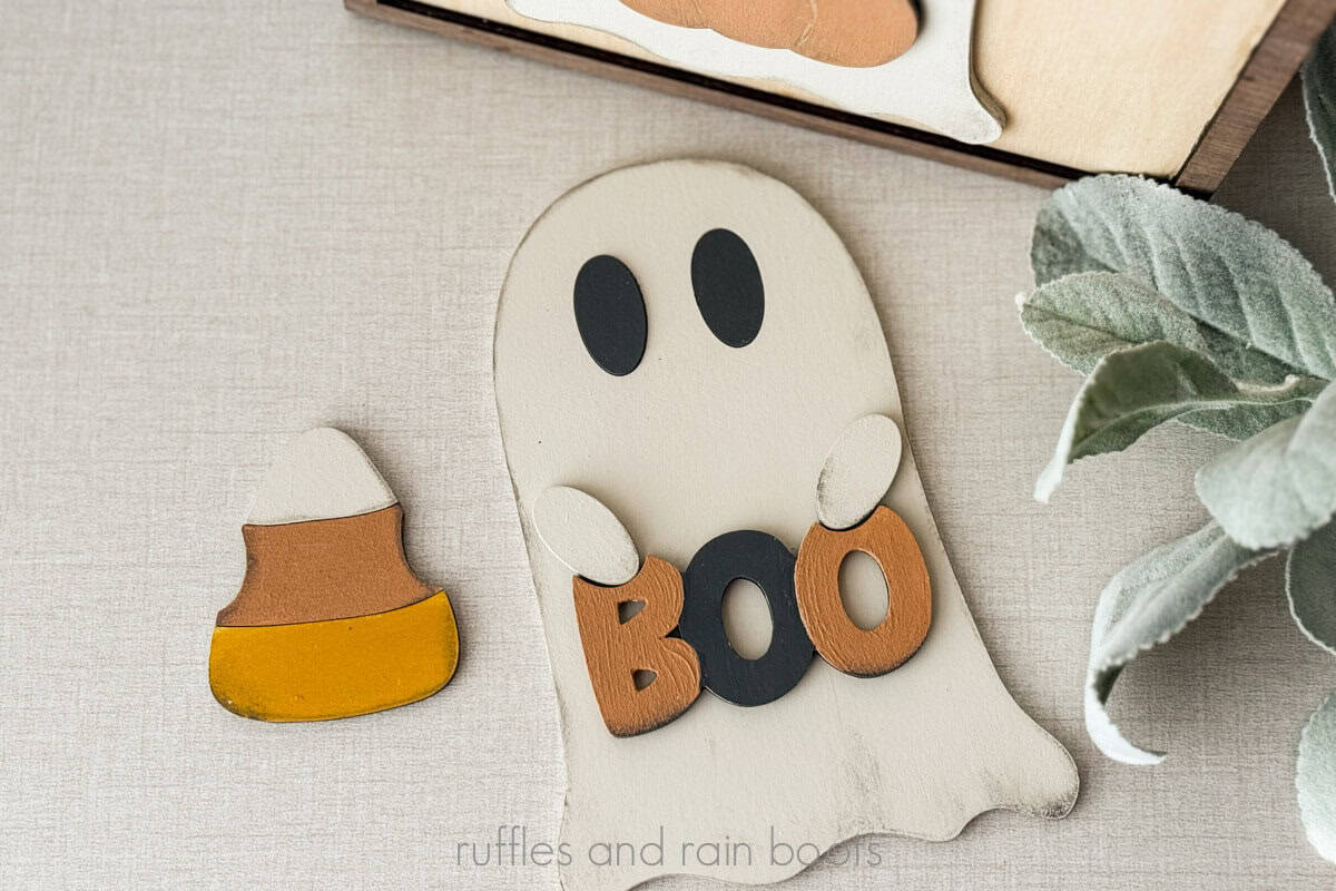Horizontal image of a laser cut ghost holding a sign which reads BOO, next to a tri color candy corn.