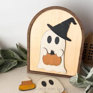 Square close up image of a Halloween ghost in a witch hat holding a pumpkin.