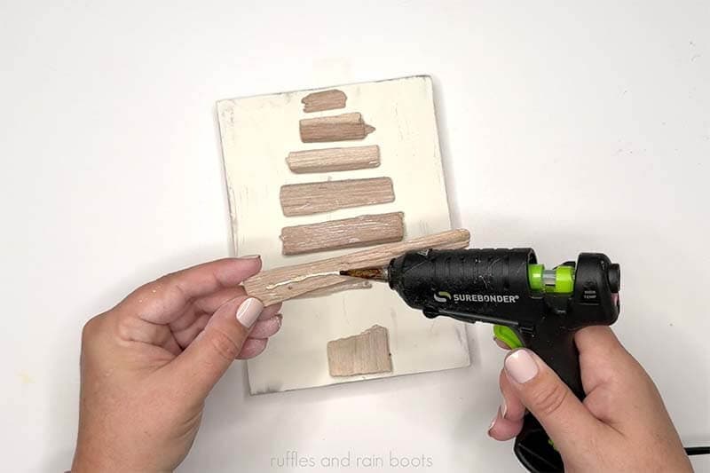 Horizontal image of a crafter holding a glue gun and attaching pieces of driftwood to a painted board.