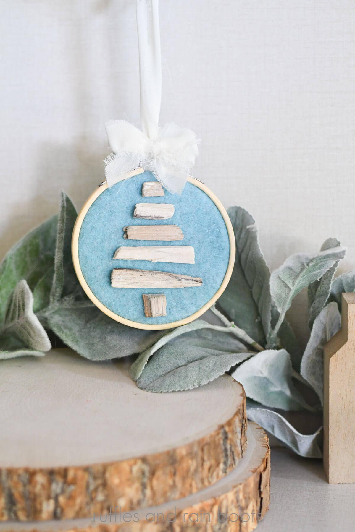 Vertical close up image of a driftwood Christmas tree ornament in an embroidery hoop with chiffon bow in front of lamb's ear and wood rounds.