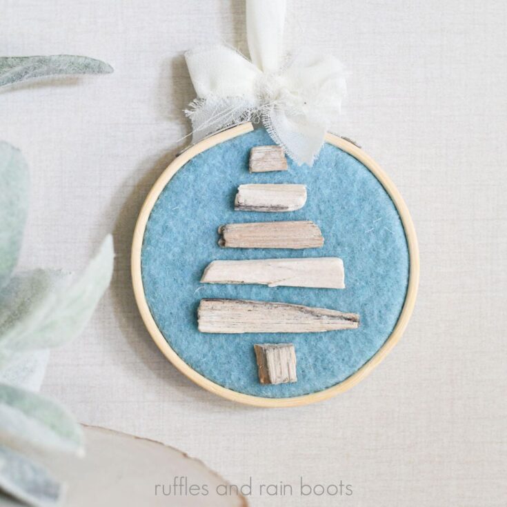Square close up image of an ocean blue felt covered embroidery hoop with a mini driftwood Christmas tree and a cream chiffon ribbon bow.