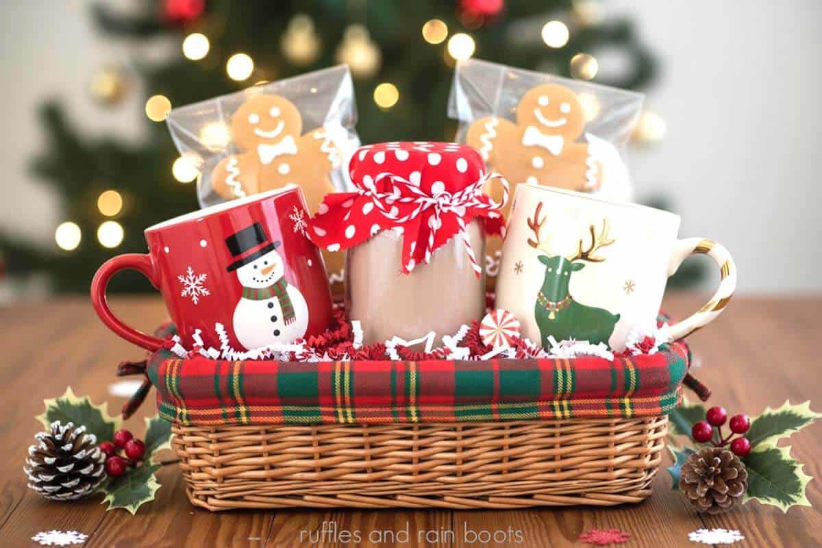 Horizontal close up image of a gift basket in a Ralph Lauren Christmas style with two holiday mugs, gingerbread cookies, and a homemade hot cocoa powder mix recipe in a jar with a fabric cover.