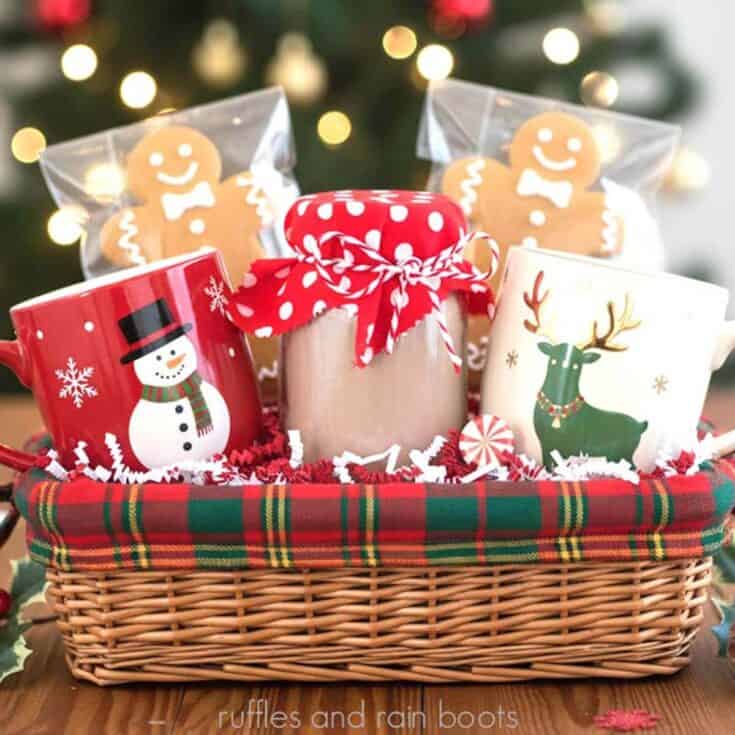 Square image of a Christmas gift basket with two holiday mugs, gingerbread cookies, and a homemade hot cocoa powder mix recipe in a jar with a fabric cover.
