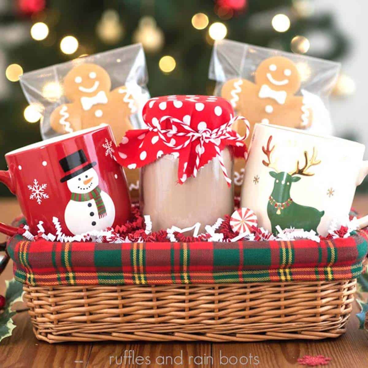 Square image of a Christmas gift basket with two holiday mugs, gingerbread cookies, and a homemade hot cocoa powder mix recipe in a jar with a fabric cover.