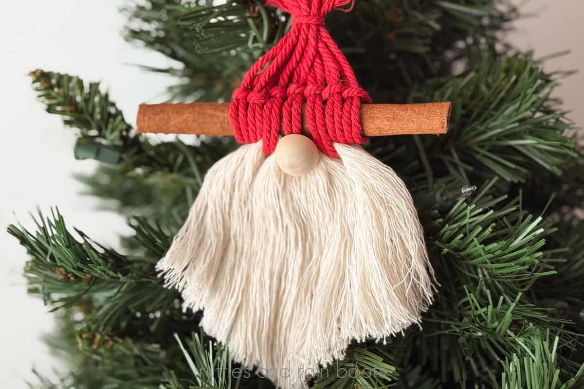 Horizontal image of a macrame gnome ornament made with cord, a wood bead, and a cinnamon stick and hung on a small holiday tree.
