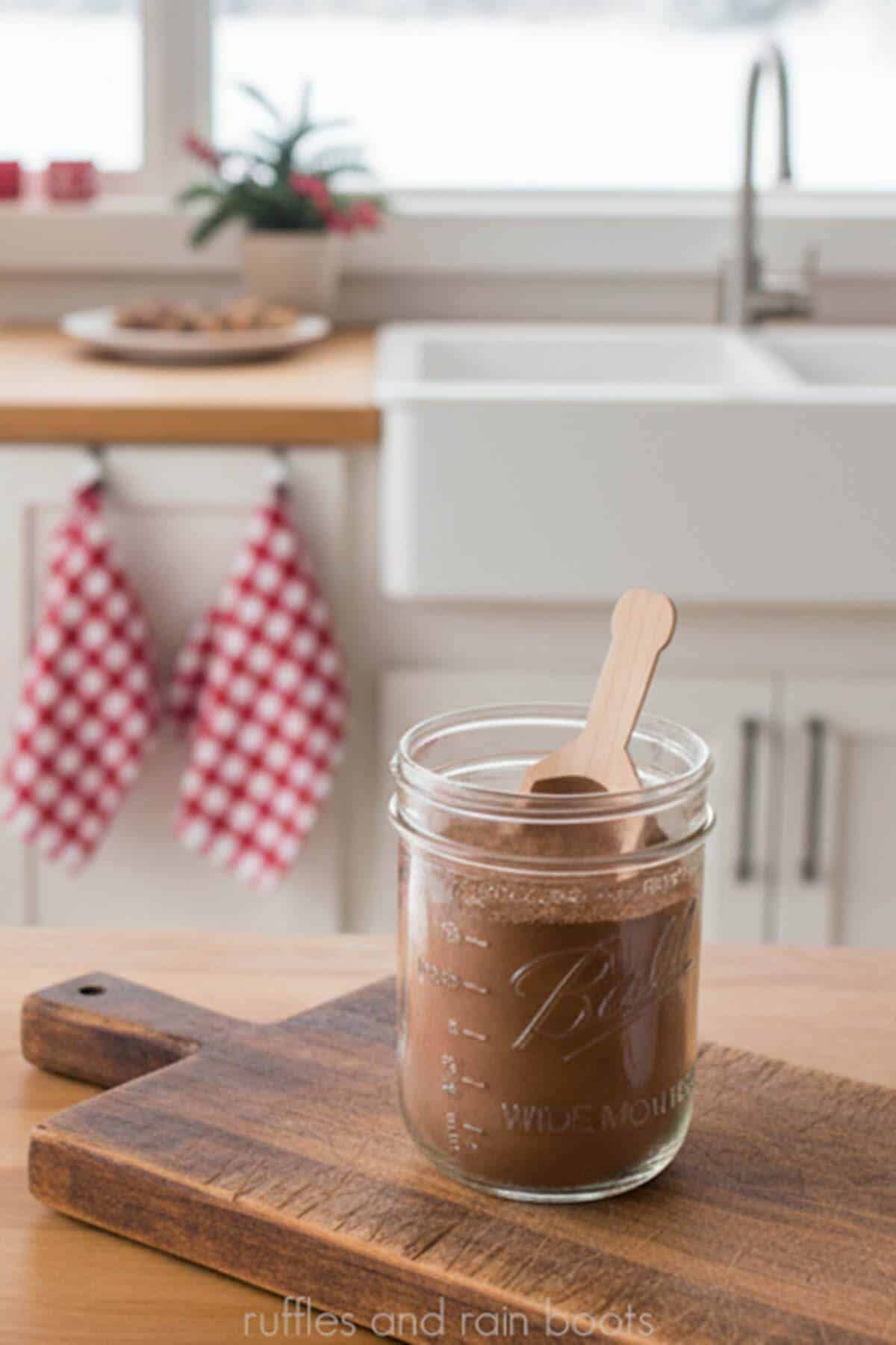 Vertical image of a mason jar full of hot cocoa mix and a wooden spoon on a wood cutting board in a white farmhouse kitchen with red checkered dish towels and a Christmas cactus.