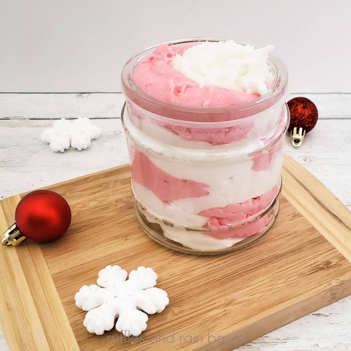 Close up square image of a white and pink whipped sugar scrub in a mason jar placed on a cutting board surrounded by snowflakes and Christmas ornaments.