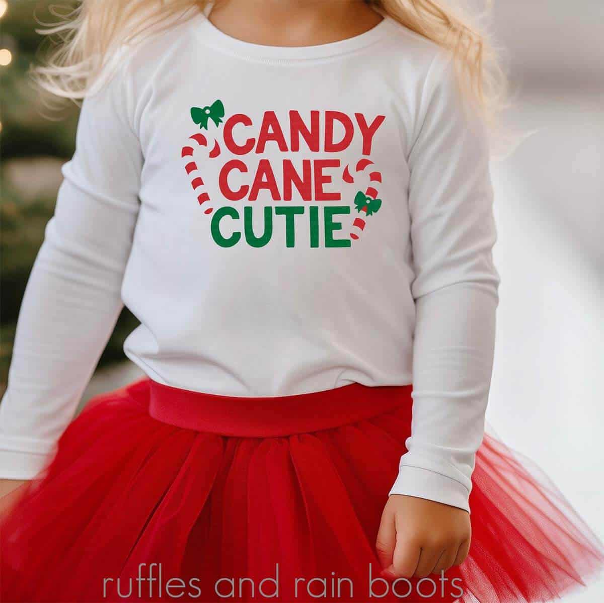 Square image of a little girl wearing a white long sleeve shirt with candy cane cutie in red, white, and green vinyl standing in front of a decorated Christmas tree.