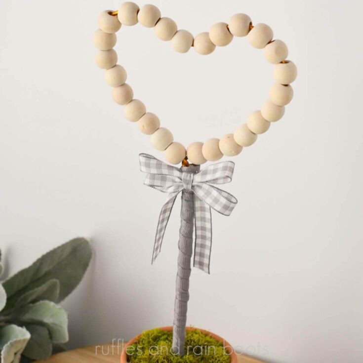 Close up square image of a wood bead heart topiary and ribbon stem in a moss topped clay pot amongst lamb's ear.
