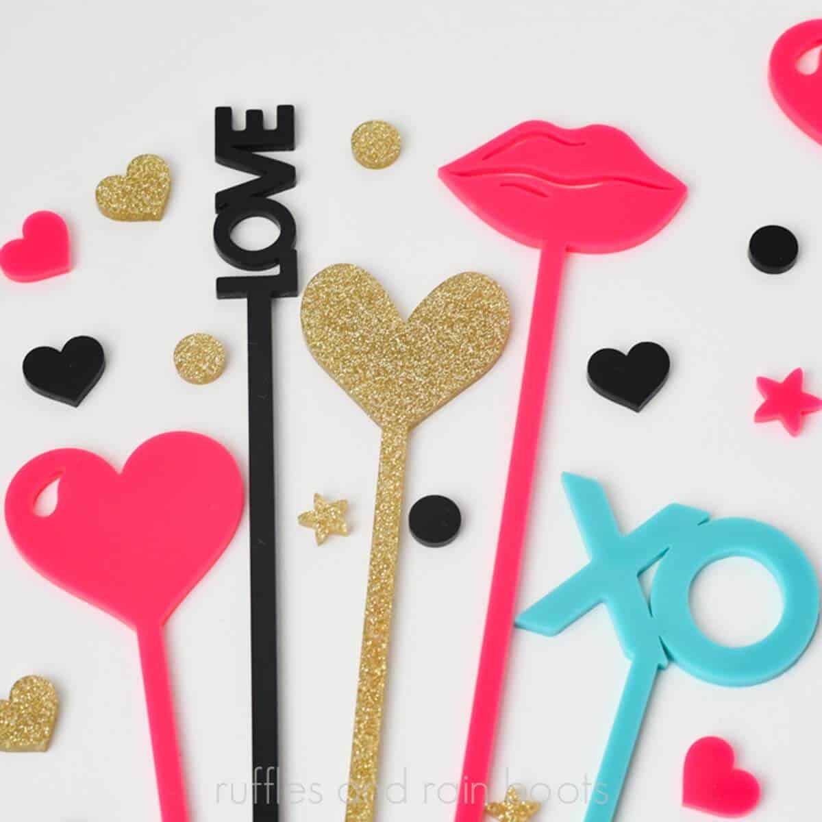 Square close up image of pink, gold, black, and teal stir sticks in a Valentine's Day theme on a white table with acrylic confetti.