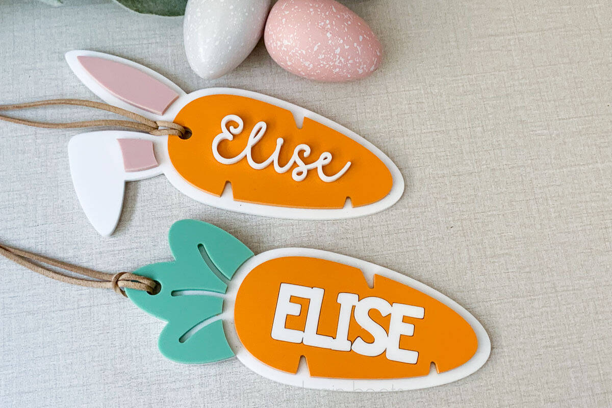 Horizontal close up image of two carrot Easter basket tags made with acrylic, placed on a linen background with faux speckled eggs.
