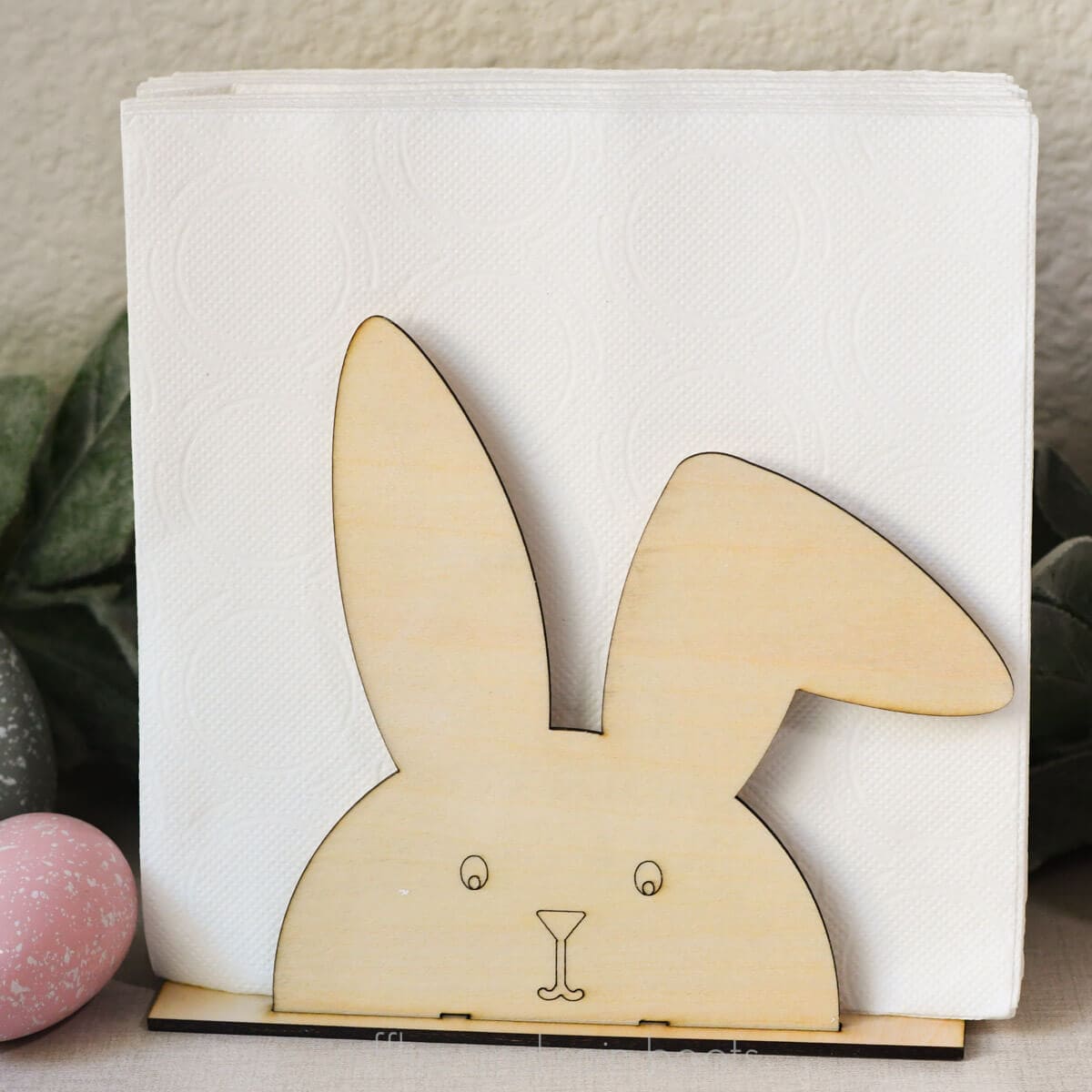Square, close up image of a wood bunny napkin holder with a cut base, two sides, and a score rabbit face holding white paper napkins.