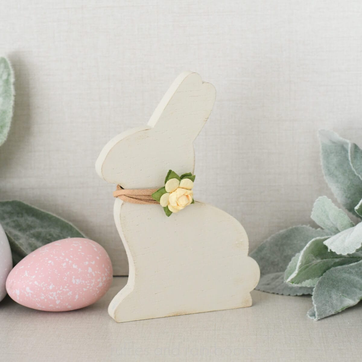 Close up square image of a white bunny with a flower at the neck in front of lamb's ear greenery and painted Easter eggs.