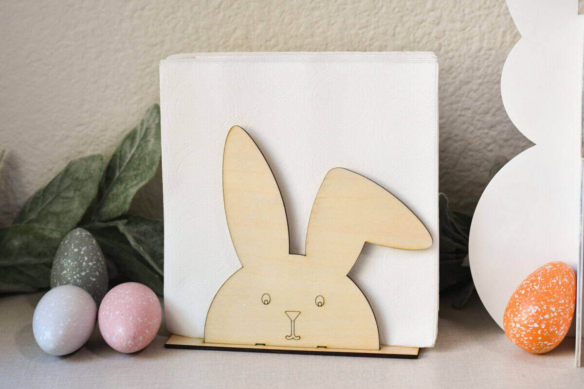 Horizontal, close up image of a wooden bunny napkin holder with white napkins nestled within greenery and colorful farmhouse Easter eggs.