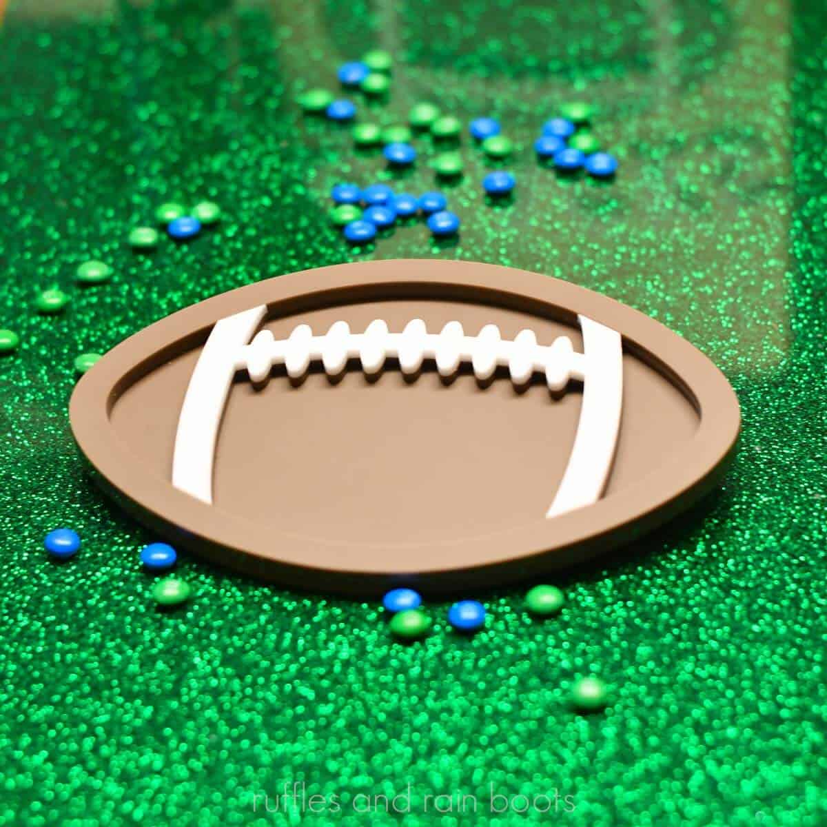 Close up square image of a layered acrylic football candy dish on a bright green glitter background with scattered mini M&Ms in blue and green.
