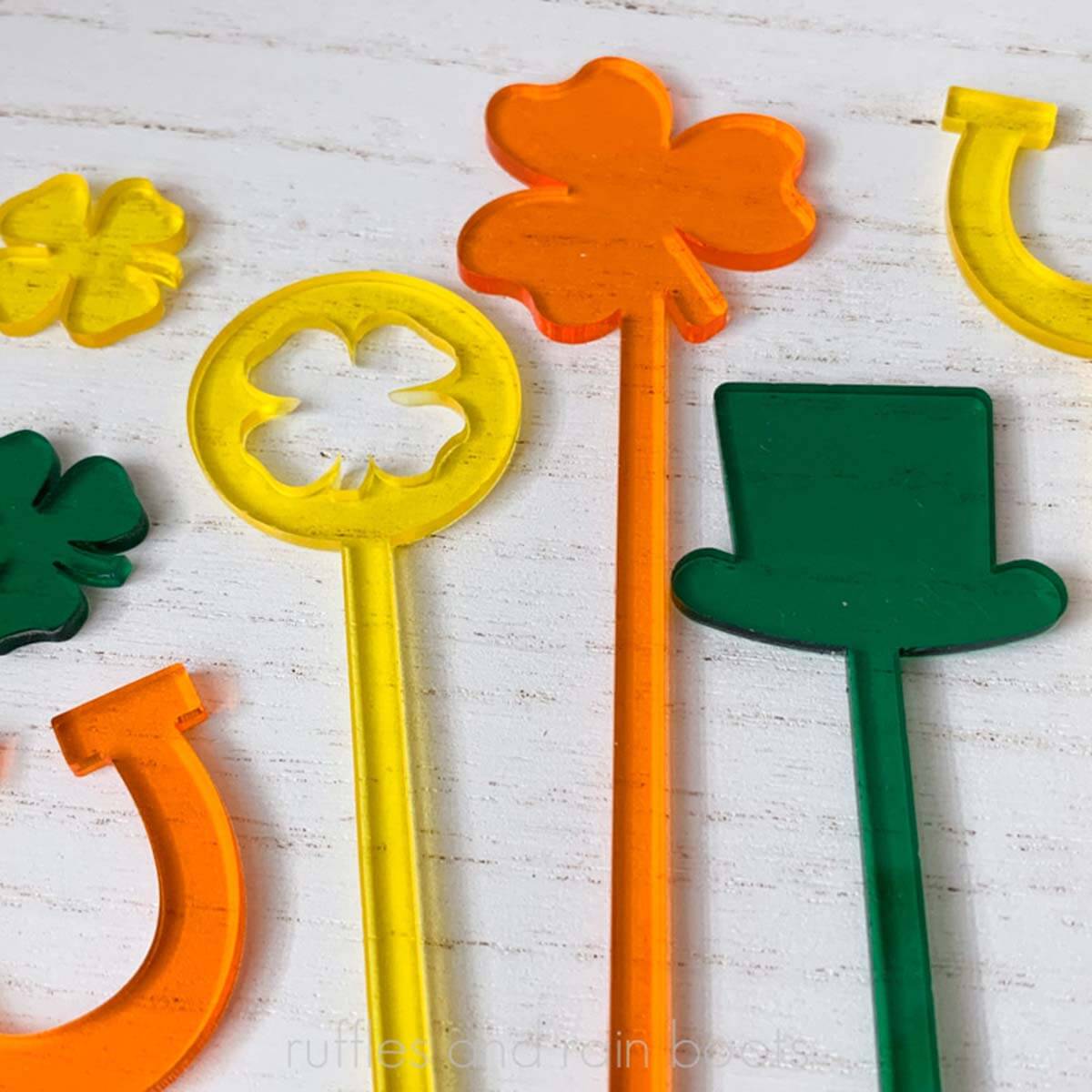 Square close up image of green, orange, and yellow translucent cast acrylic St Patricks Day themed drink stir sticks on wood table.