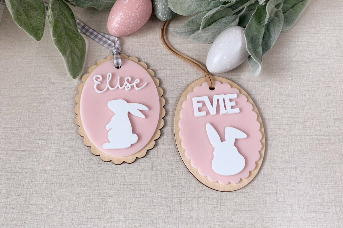 Horizontal image of two scallop edge Easter basket tags made of pink and white acrylic with basswood backers on a linen background with eggs and greenery.