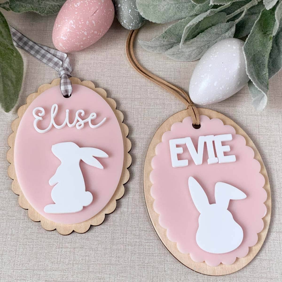 Square, close up image of two scallop edge Easter basket tags in pink and white acrylic on wood backers with names and bunnies.