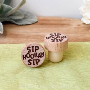 Square, close up image of two laser engraved wine bottle stoppers on a green organza and neutral background.