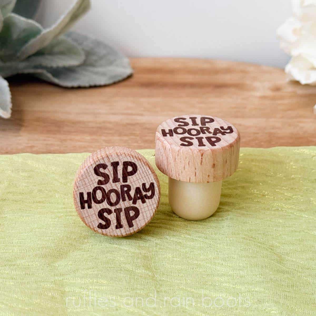 Square, close up image of two laser engraved wine bottle stoppers on a green organza and neutral background.