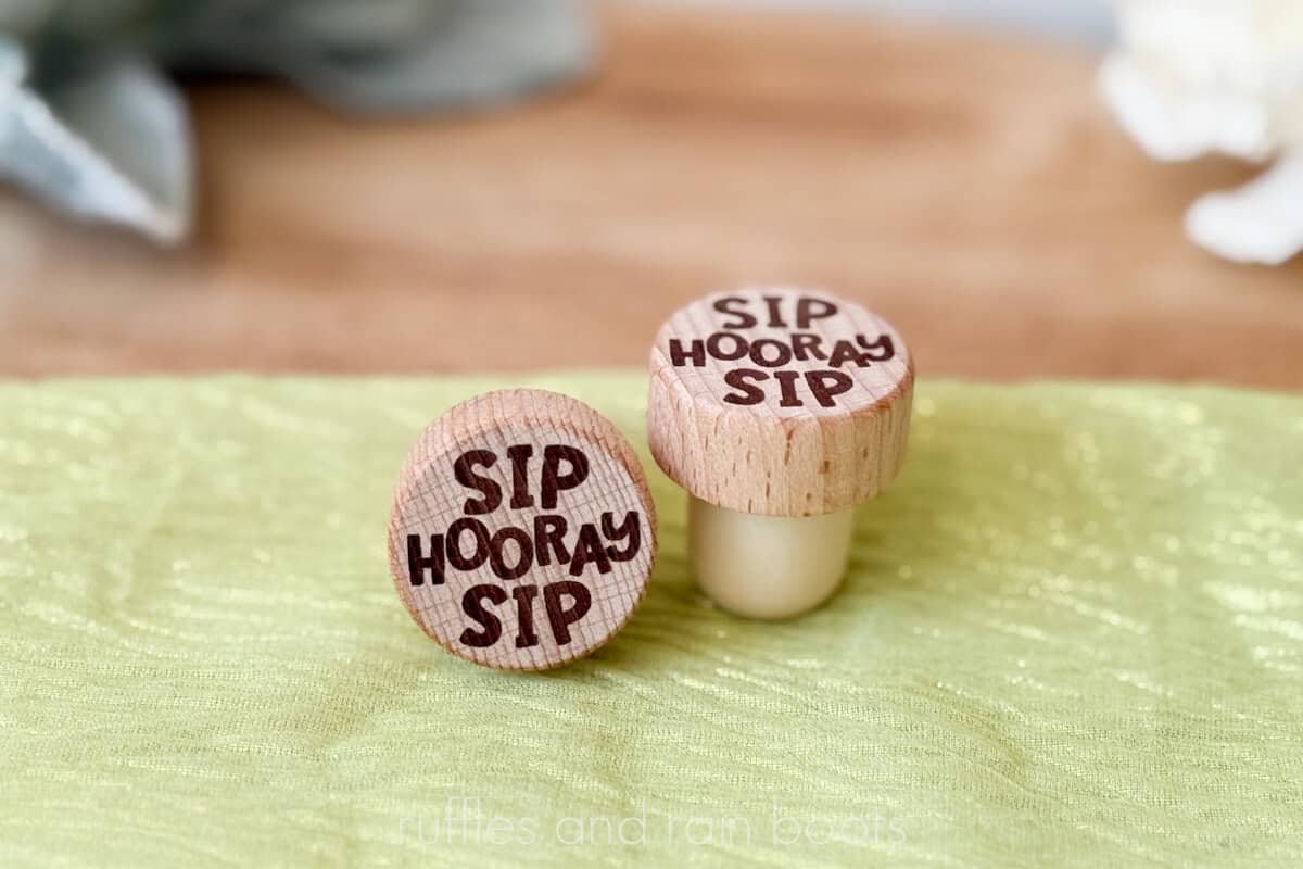 Horizontal image of two wooden laser engraved wine bottle stoppers which read sip sip hooray on a green organza and a neutral, natural background.