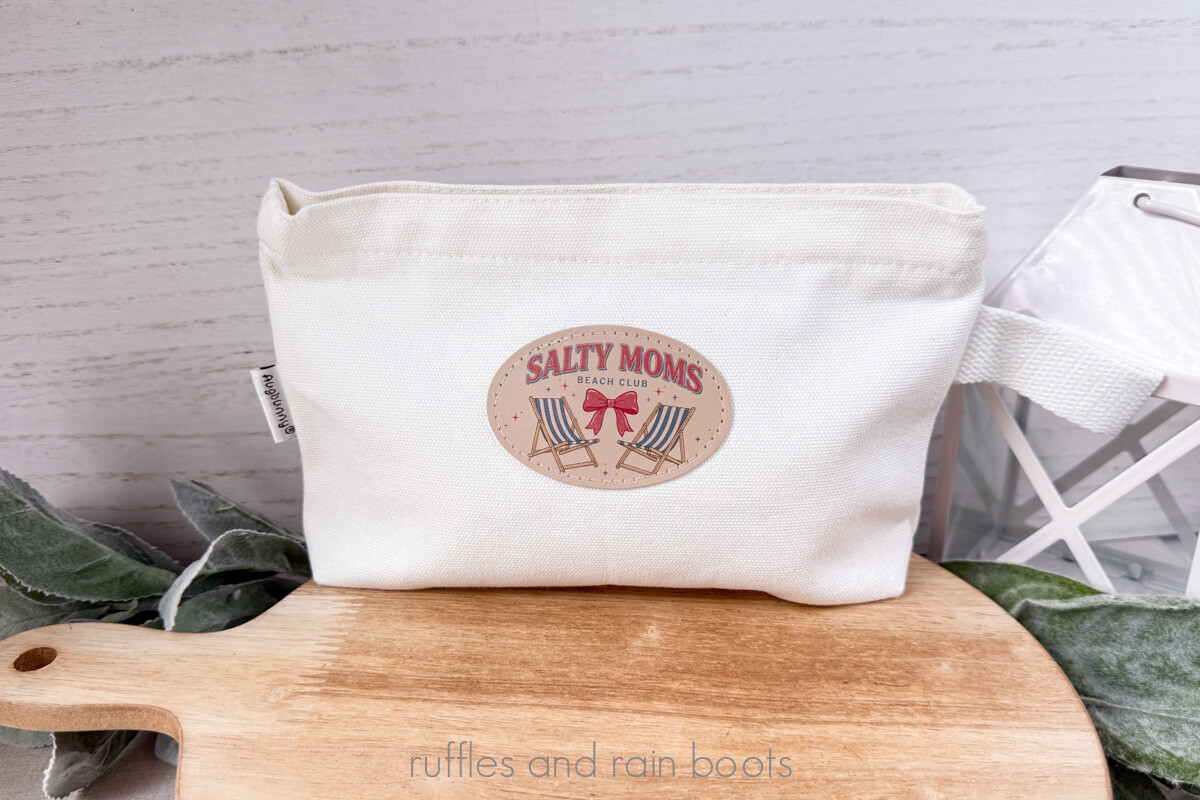 Horizontal image of a white make up bag with tab on a wooden cutting board showing a heat pressed printed leather patch, created by the xTool M2 printer and laser machine.