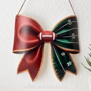Square, close up image of a football bow sign with faux leather on one side, a velvet ribbon mimicking the football field's yard line, and a football in the center hanging against a plain wall.