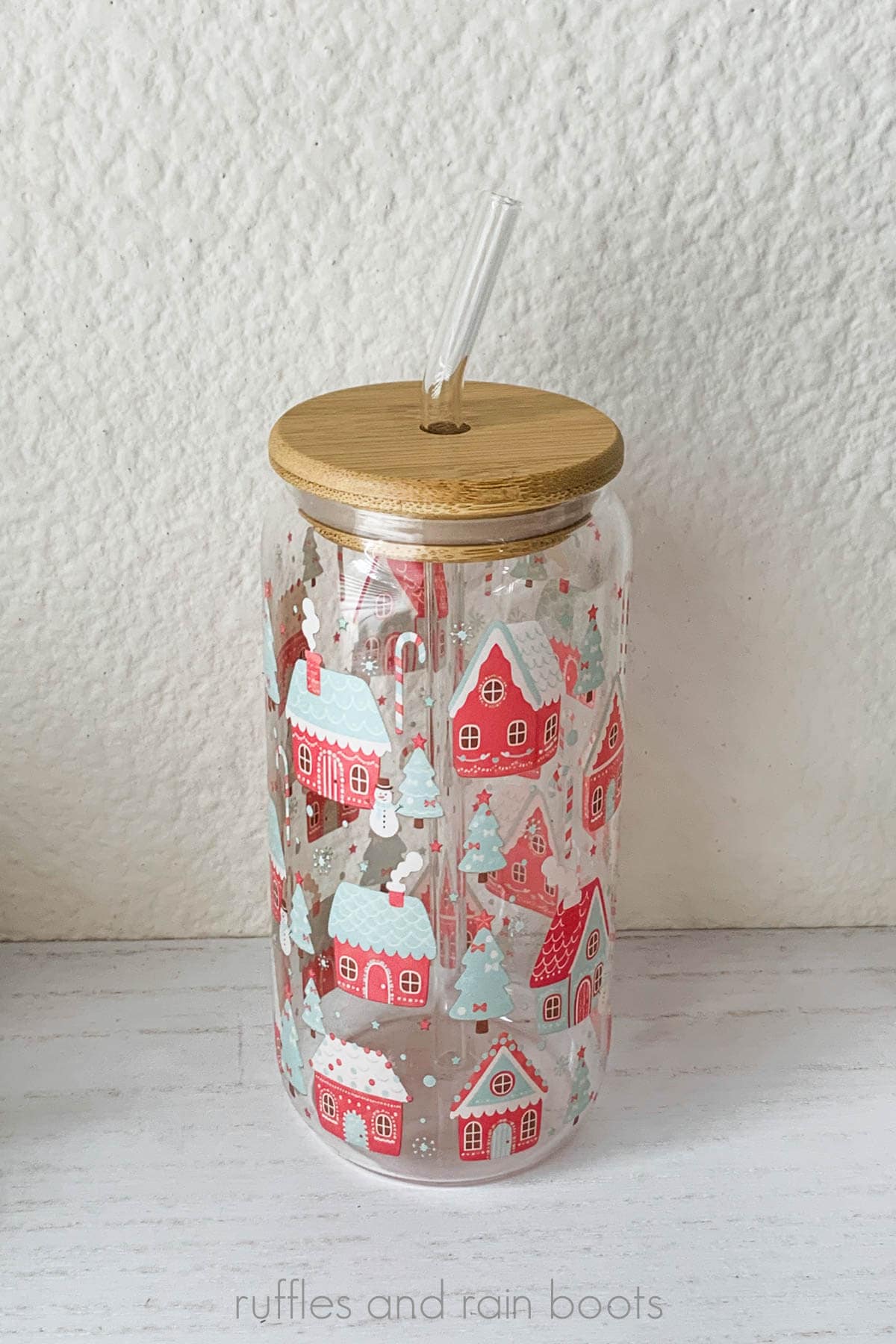 Vertical close up image of a glass drinking tumbler with a bamboo lid and glass straw printed with a pastel Christmas theme on a plain white board against a light beige wall.