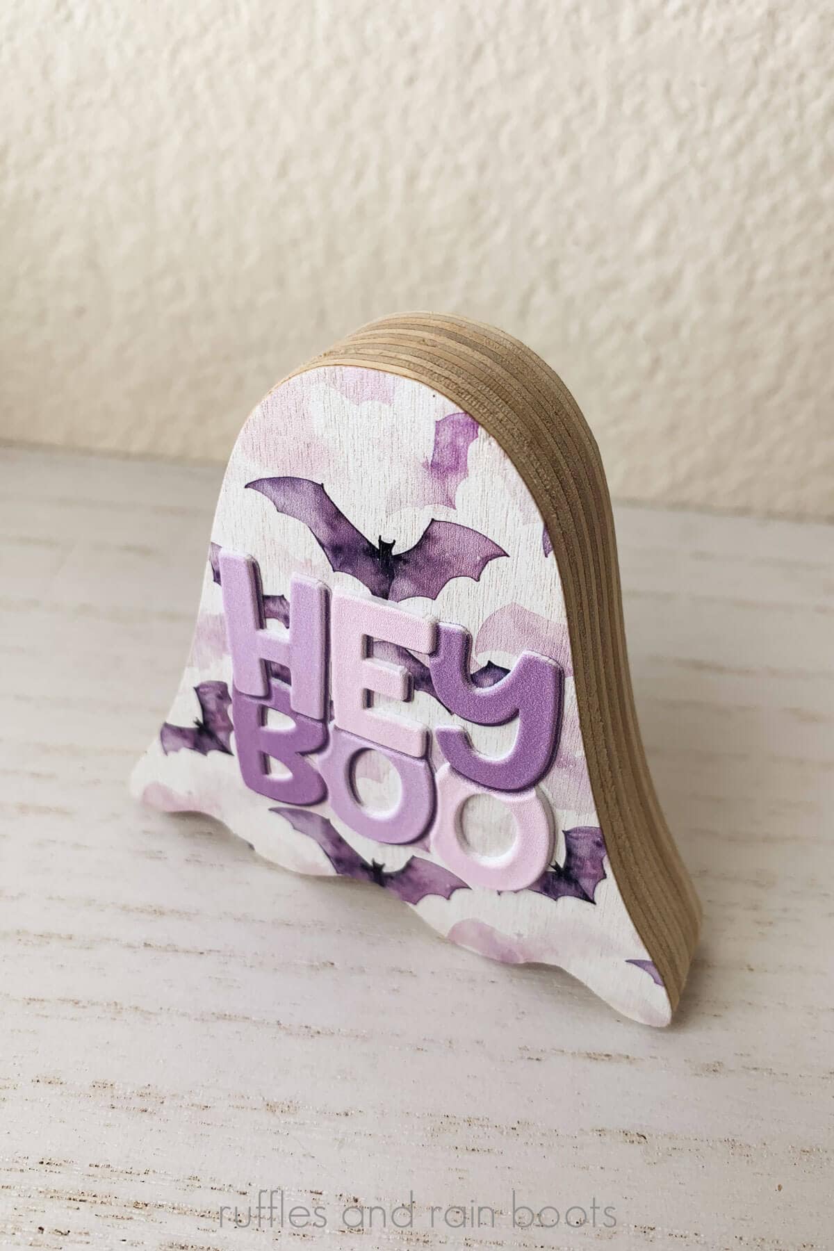 Close up vertical image of a wood ghost printed with a watercolor pink and purple background and raised texture text which reads hey boo in pinks and purples.